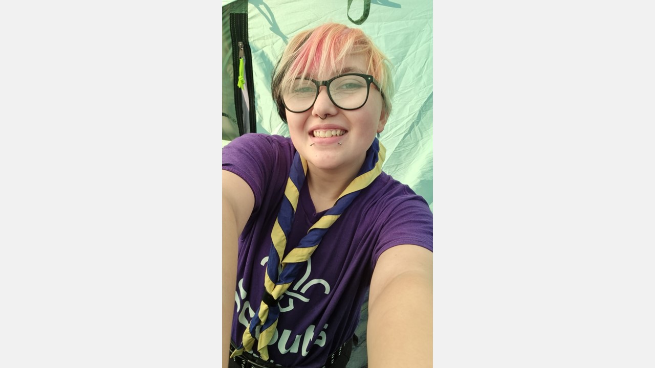A young person wearing glasses, a necker and a purple Scouts t-shirt