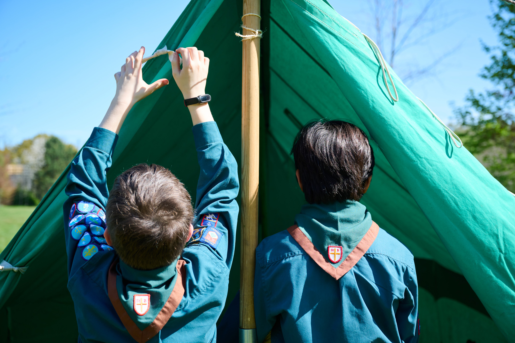 The image shows the back of two Scouts who are facing a tent. They're both in uniform with neckers on. The Scout to the left has brown hair and is holding their hands up and holding part of the green tent.