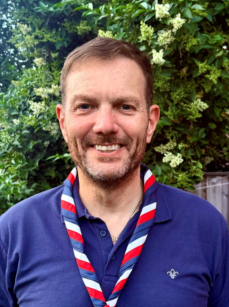 A headshot of a man wearing a blue polo shirt and a UK Scouts necker