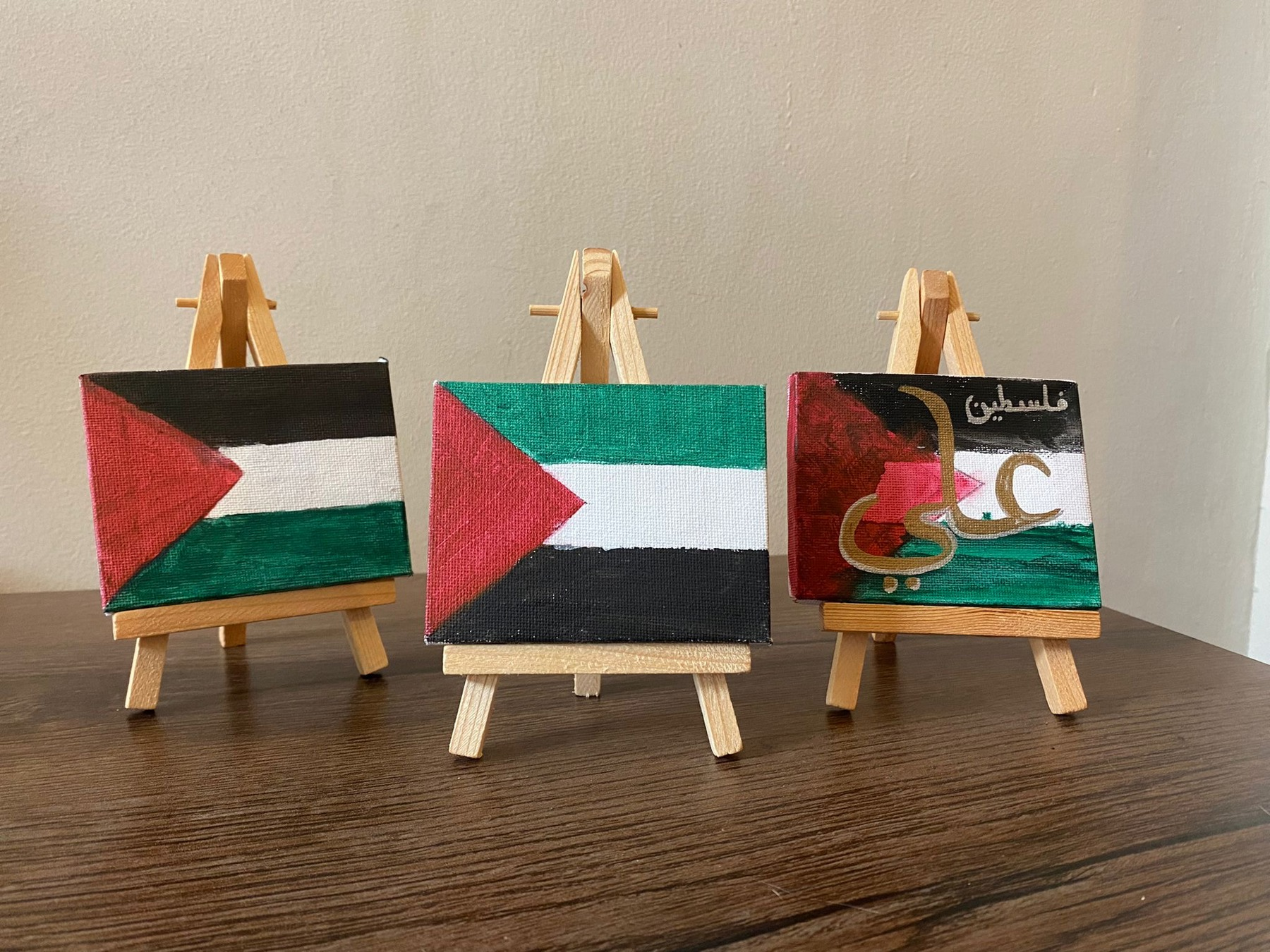 The image shows Palestine flags painted on small canvases on wooden stands. There are three in total on top of a wooden table in front of a wall.