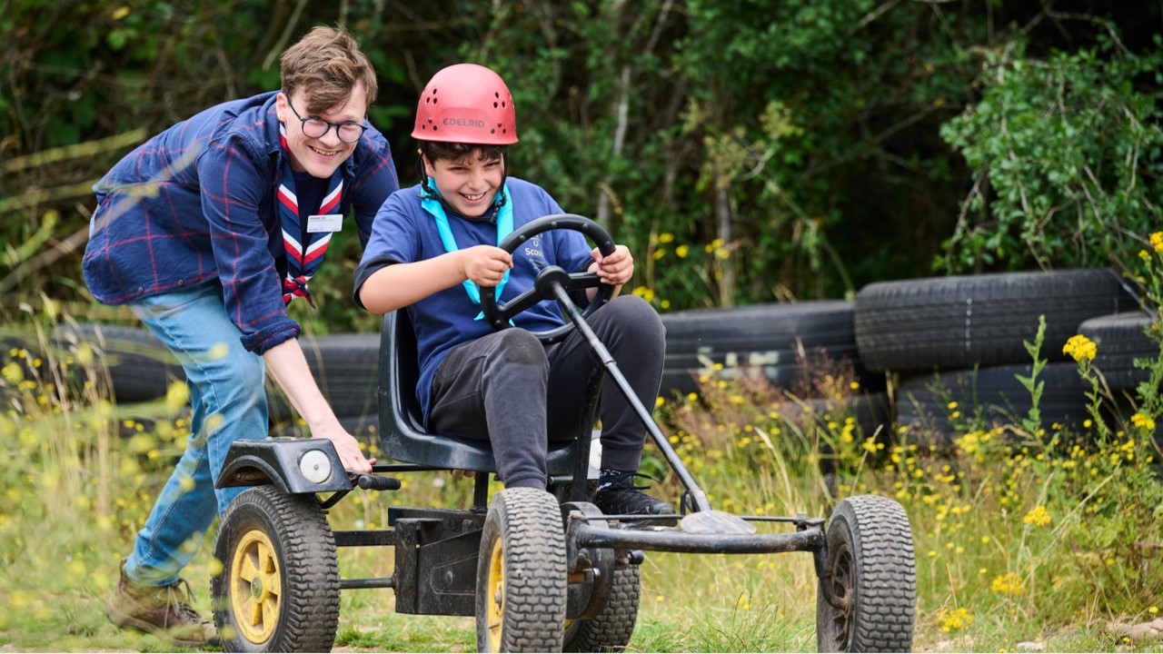A young person sits in a go-kart smiling, while a volunteer pushes the car from behind.