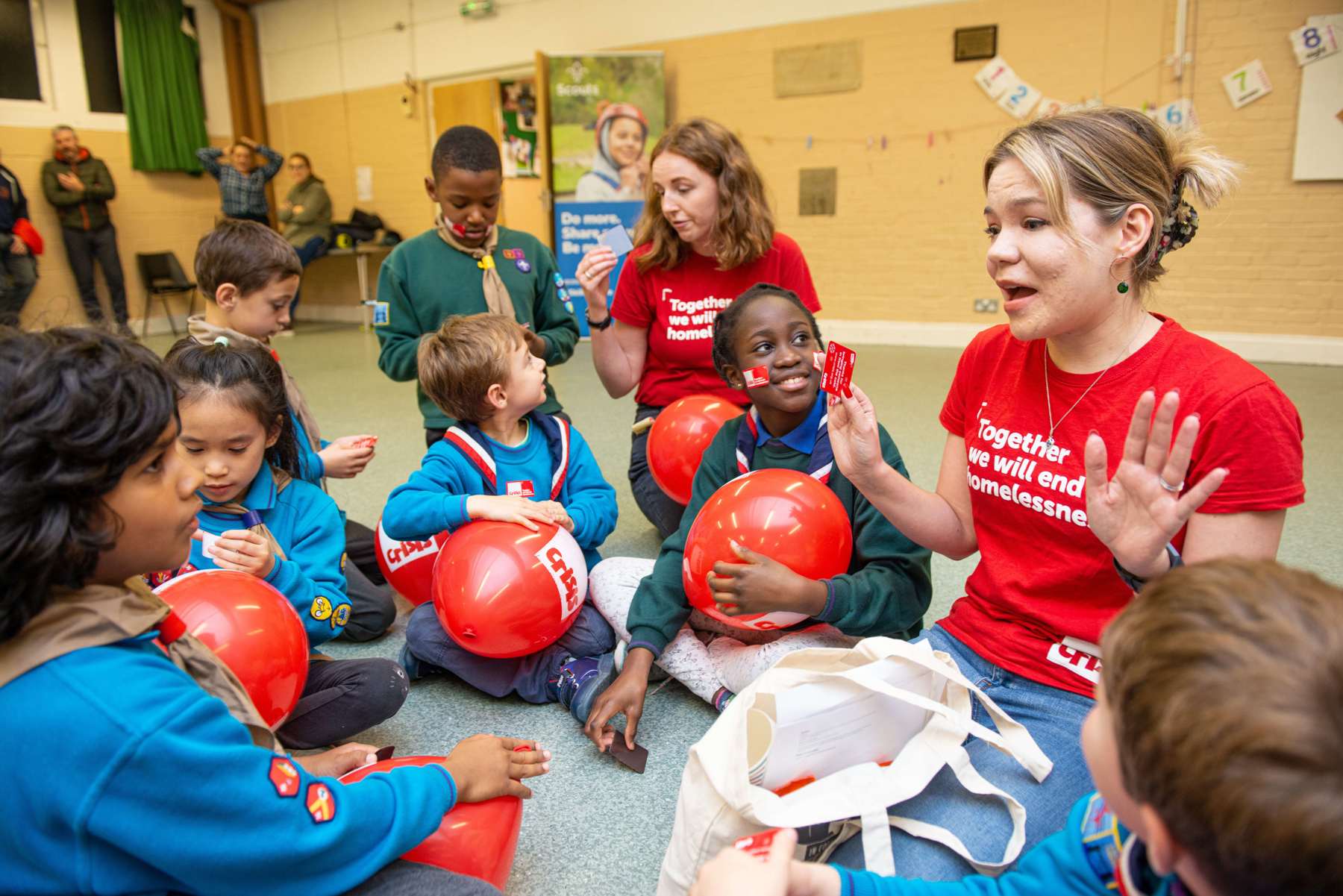 Beavers and Cubs sit on the floor looking at and listening to a person from Crisis.