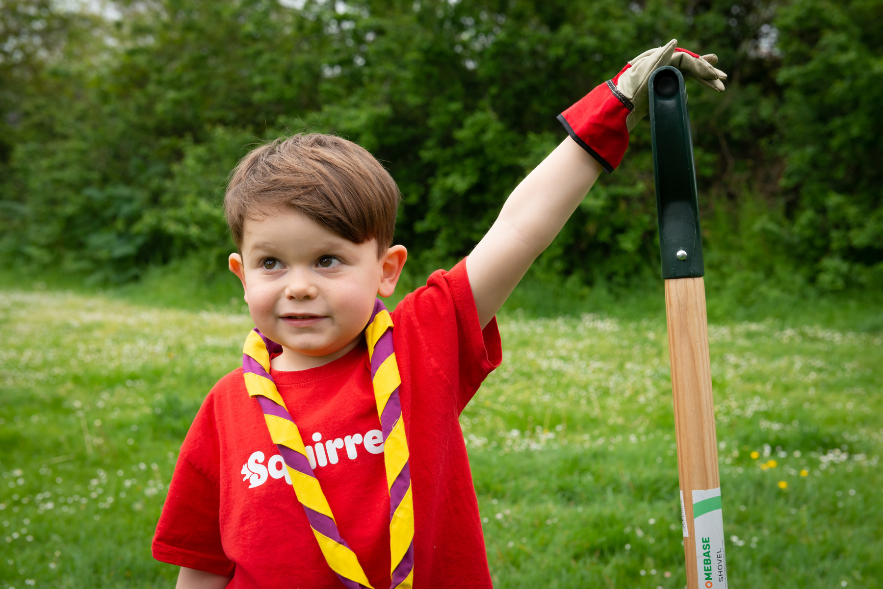 A Squirrel is standing on grass wearing a red Squirrels t-shirt and a red and yellow necker. Their left hand has a glove on and is holding the top of a spade.