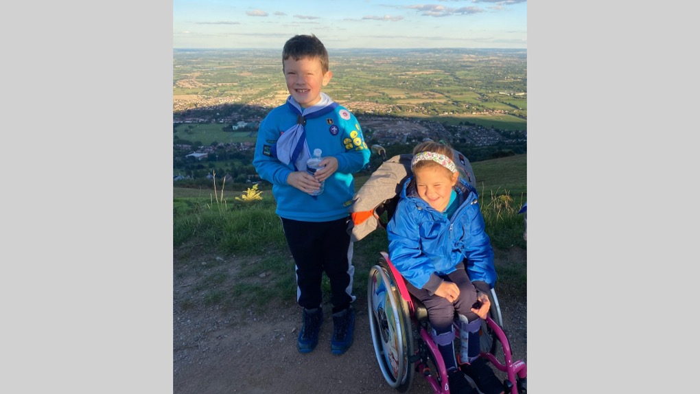 The image shows Sophie in her wheelchair and Beaver uniform outside on a hill. She's smiling and looking towards the ground with the sun on her face. Her brother is stood to her left, also in Beaver uniform and is smiling at the camera while holding a plastic water bottle in both his hands.