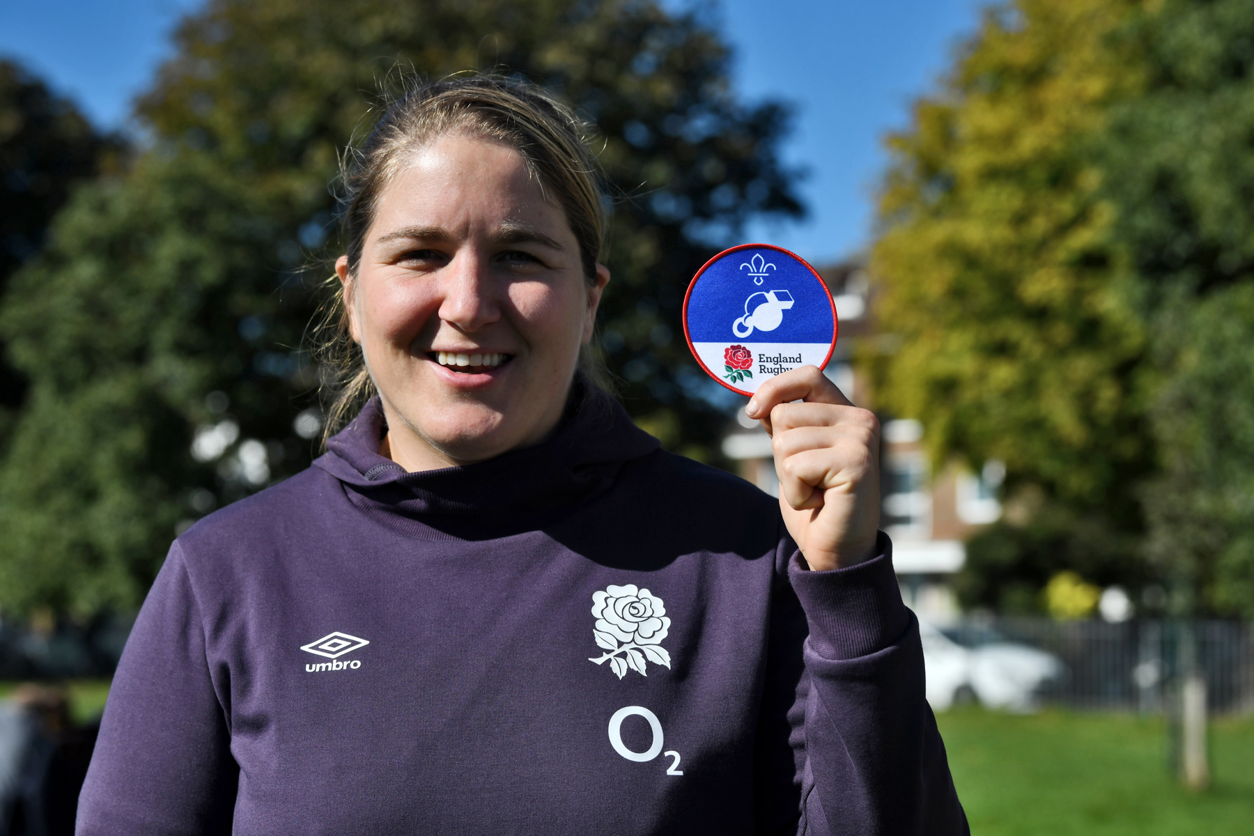 The image shows Poppy Cleall, England International, holding up our England Rugby (RFU) supported badge. She's holding the badge up next to her left ear and is wearing a grey England Rugby hoodie while smiling at the camera.