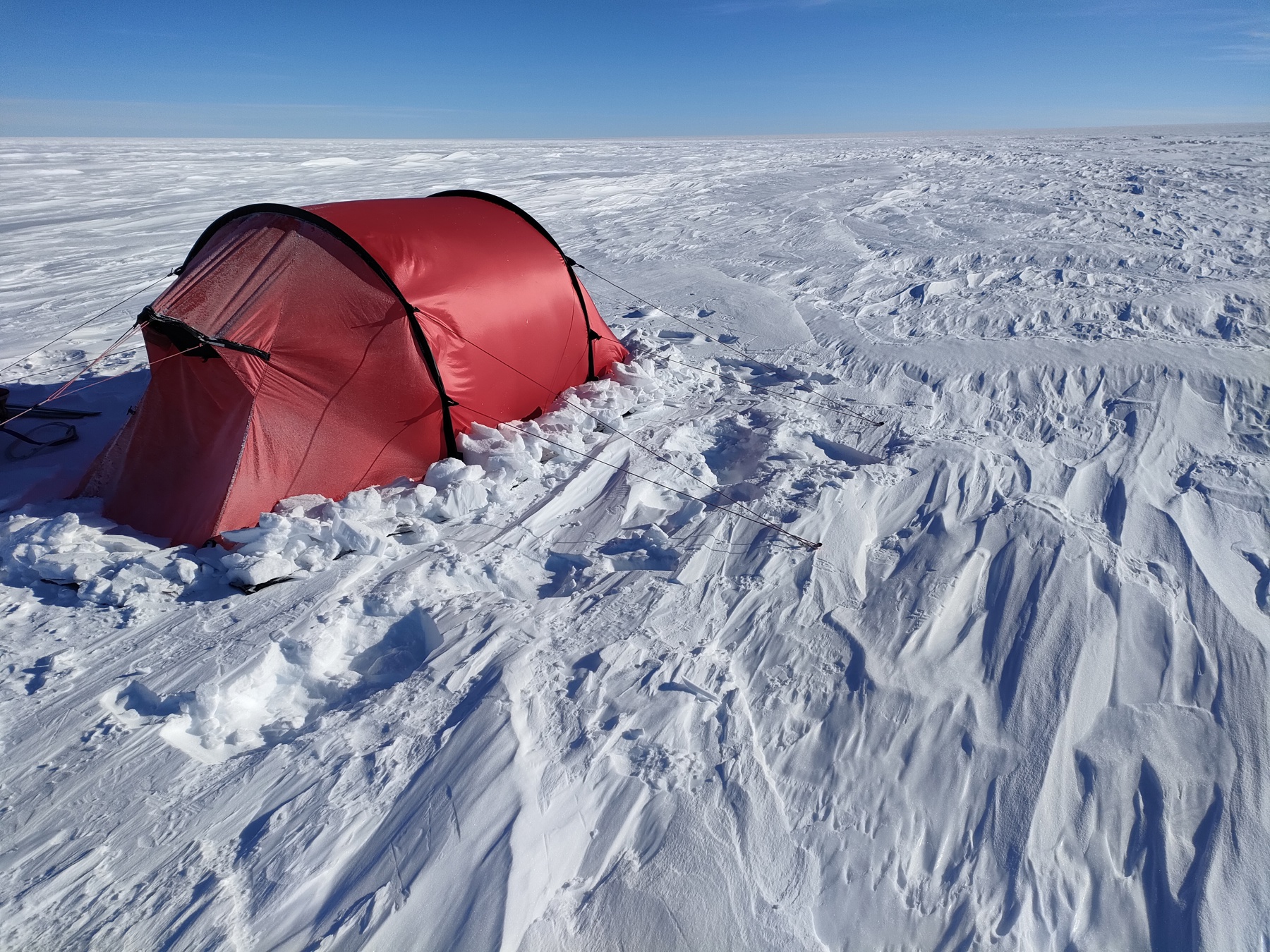 The image shows a red pitched tent on snow. The tent is to the left of the image and there is nothing but snow surrounding it. The sky behind the tent is bright blue.