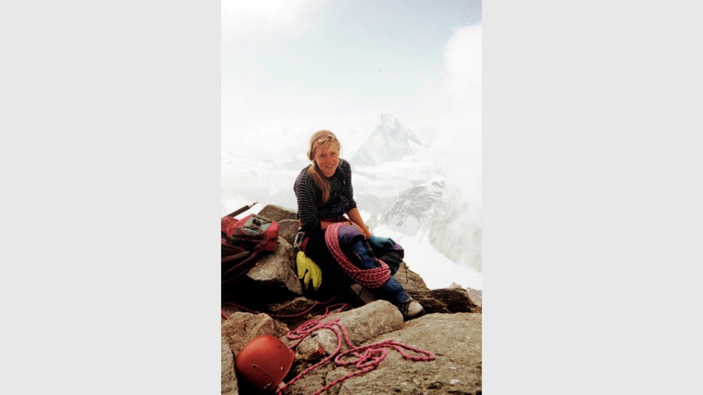 The image shows Karen Darke climbing in the Swiss alps. She's sat on the top of a mountain and looking at the camera with rope draped around her, along with a backpack and helmet. There are snowy mountains behind her.