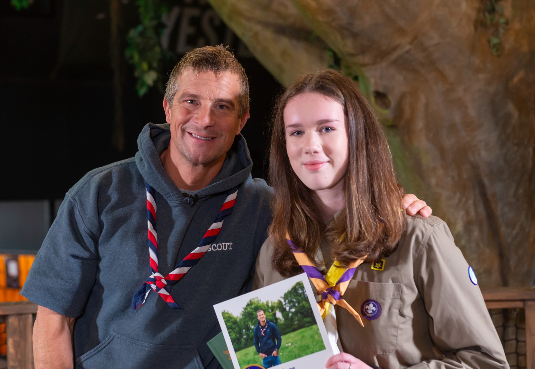 This image shows Chief Scout Bear Grylls standing next to Hannah, a teenage girl with long brown hair, who's holding up a certificate from Bear Grylls. Hannah wears a purple and orange necker, and a brown shirt with a Scout badge on. They're both smiling for the camera.