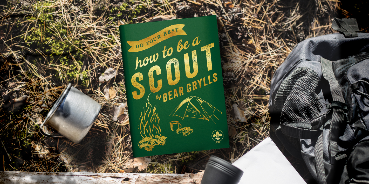 The image shows Bear Grylls' latest book, Do Your Best: How to be a Scout. The book is laid on soil ground with a metal tin pot to the left of the book, and a grey rucksack to the right.