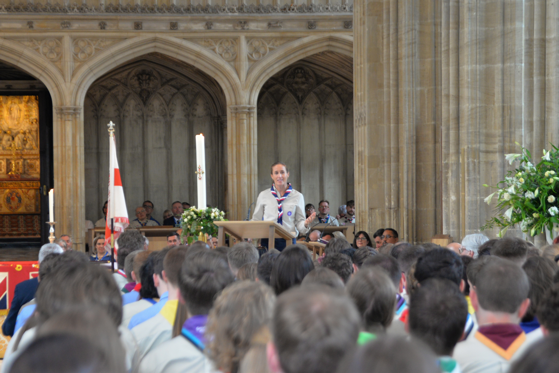 The image shows Helen Glover standing at a podium in St George's Chapel in Windsor. She is wearing a beige Scout blouse and red white and navy necker. In the foreground are the backs of people's heads as they look forward at Helen giving her address.