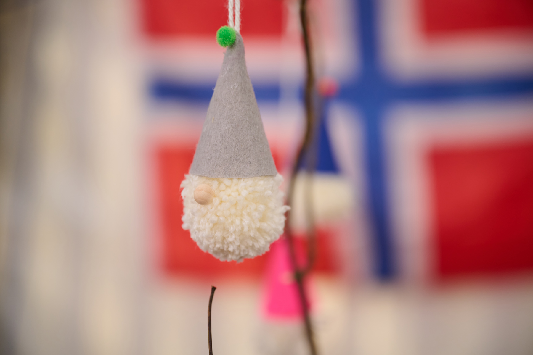 A hand holding a nisse, which is a pom pom with a wooden bead nose and a grey felt hat, with green pom pom on the top. The Norwegian flag is in the background.