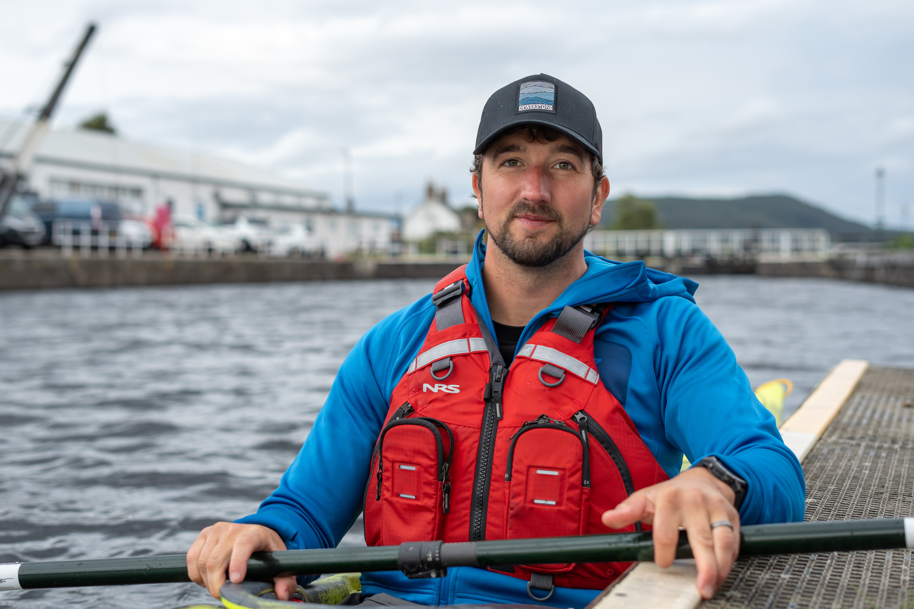 Darren is sat in a kayak on a body of water. He's looking at the camera and is wearing a cap, blue jacket and red life jacket.