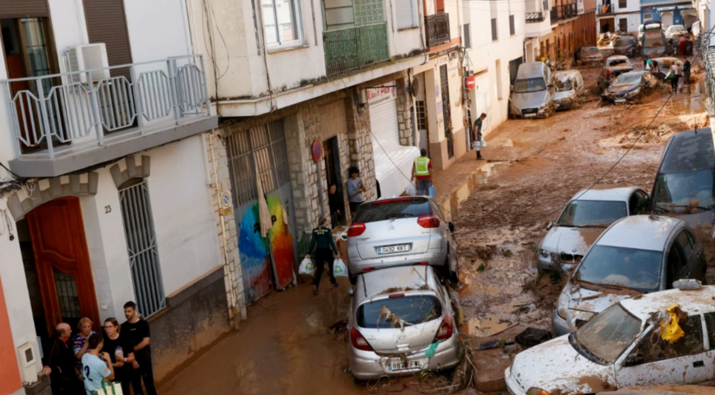 The image shows the devastating impact of floods in Spain. There are cards covered in mud and raise off the ground and destroyed, with the road covered in thick mud. To the left are apartment buildings with people gathered in the streets.