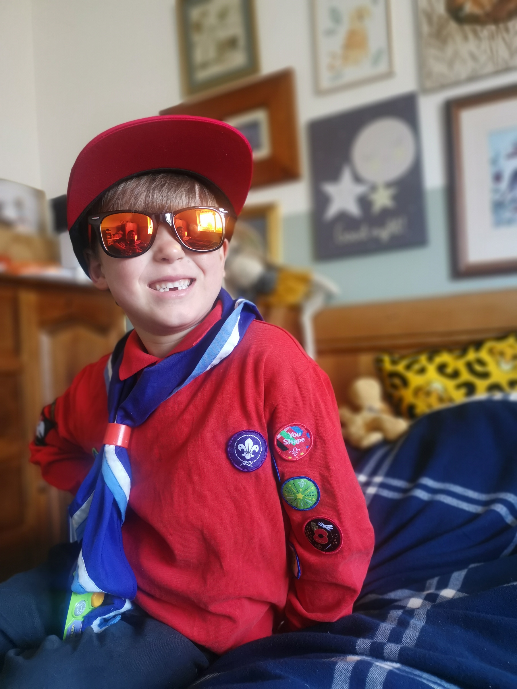 Edgar has brown hair and is sat on a bed with blue bedding. He's looking to the right. He is in a Squirrel uniform and purple necker. He is smiling and wearing sunglasses. There's lots of awards and frames on the wall behind him.