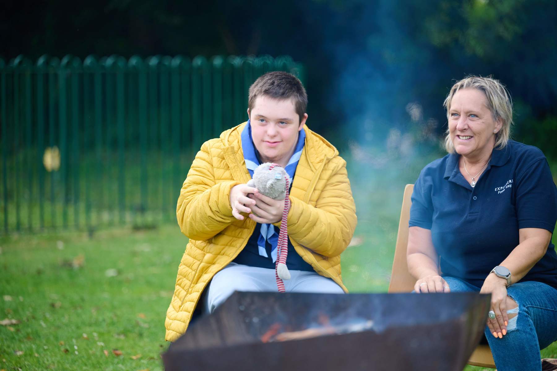 A young person holds a soft toy while sat at a campfire with a volunteer.