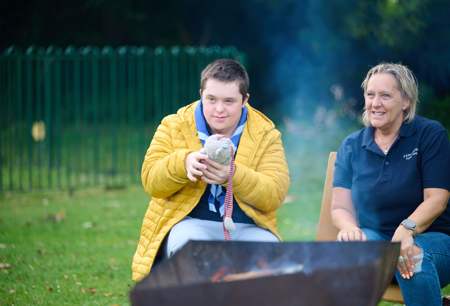 A young person holds a soft toy while sat at a campfire with a volunteer.