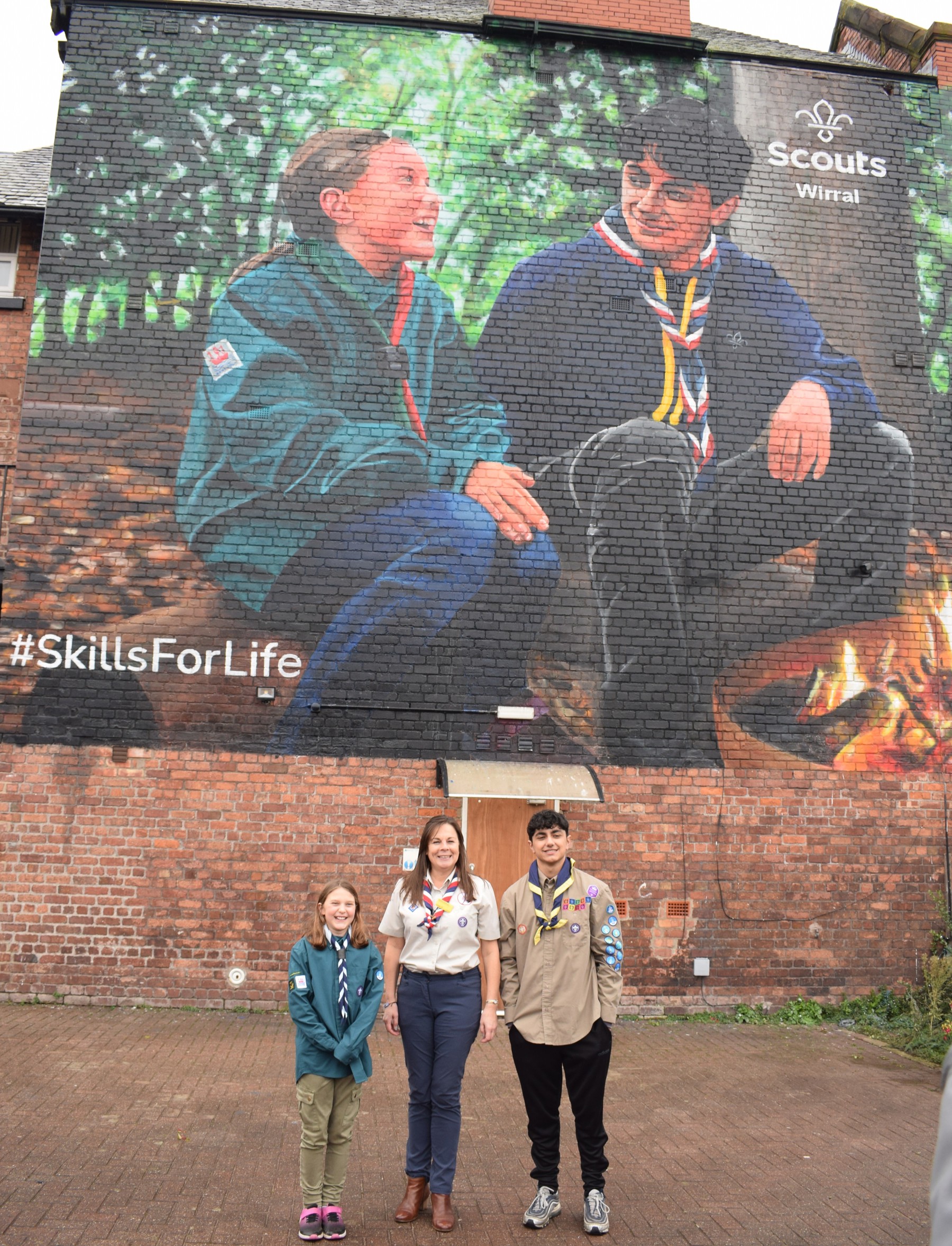 Chief Commissioner for England, Liz Henderson, stands in between Beatrice and Jack, the two Scouts featured in the Wirral Scouts mural. They're stood in front of the mural in Scout uniform and are smiling at the camera.