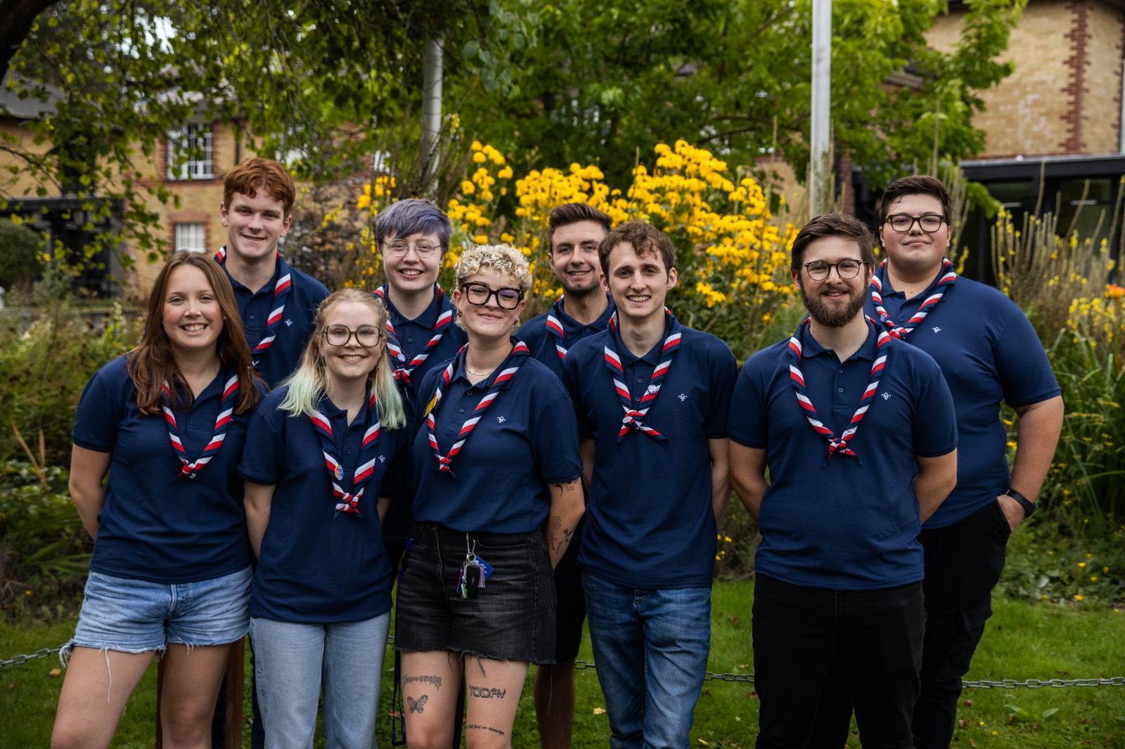 The image shows Scouts' Youth Advocates stood on grass with bushes and flowers behind them. The group of 9 are all wearing navy Scouts polo tops with red white and navy neckers. They're stood as a group with their hands behind their backs and they're smiling at the camera.