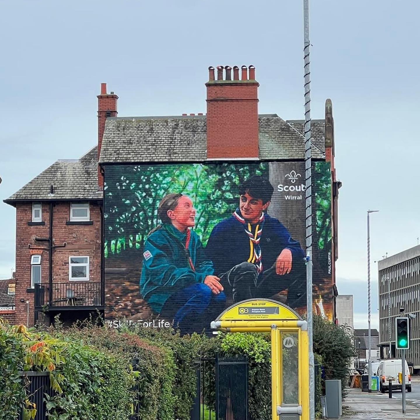 The image shows the Wirral Scouts mural, painted on the side of a brick building. The photo has been taken from the street, and there is a yellow bus stop and lamp post next to a hedge in front of the mural. The photo aims to show the size of the mural to someone walking down the street.