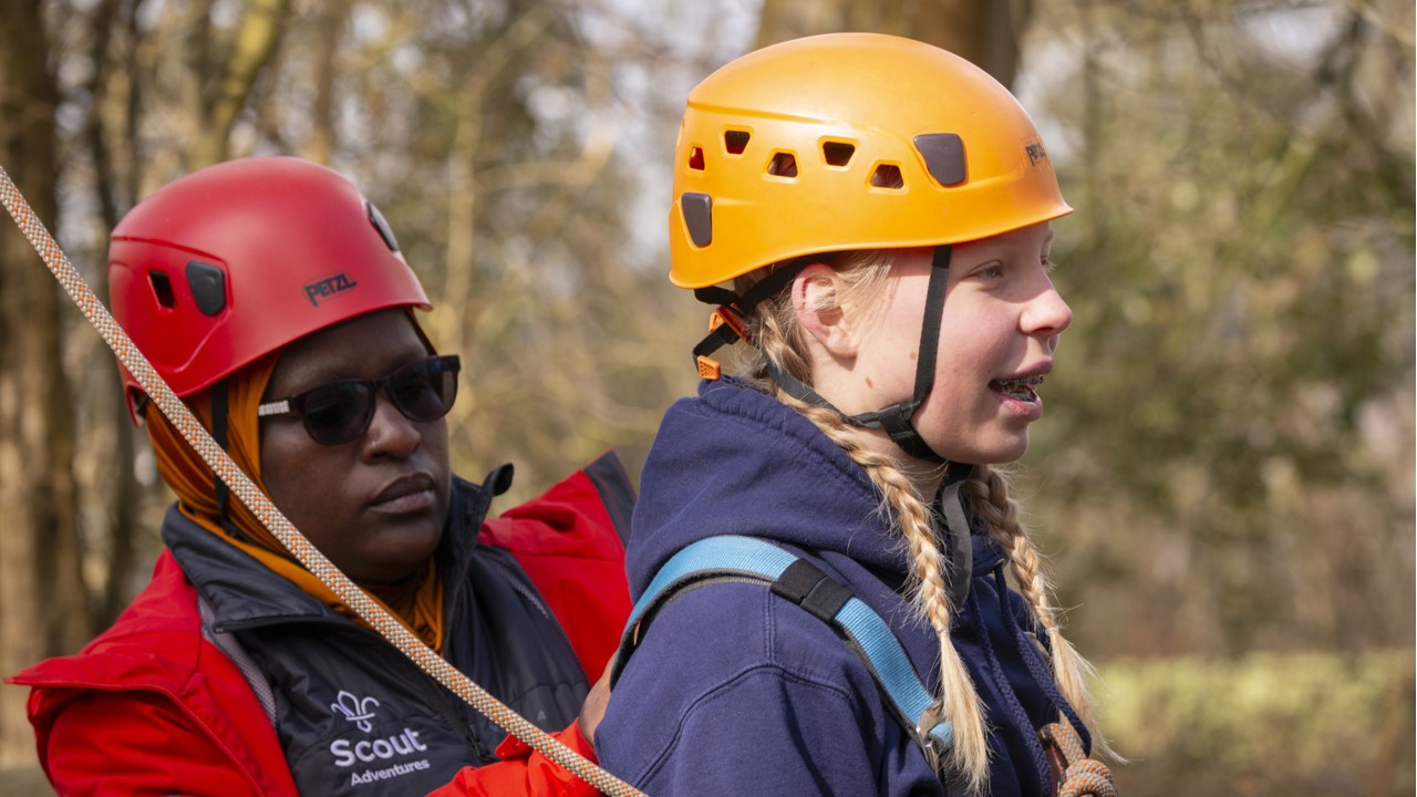 A young person, wearing a yellow helmet, looks forward while a volunteer, wearing a red helmet, secures their harness.