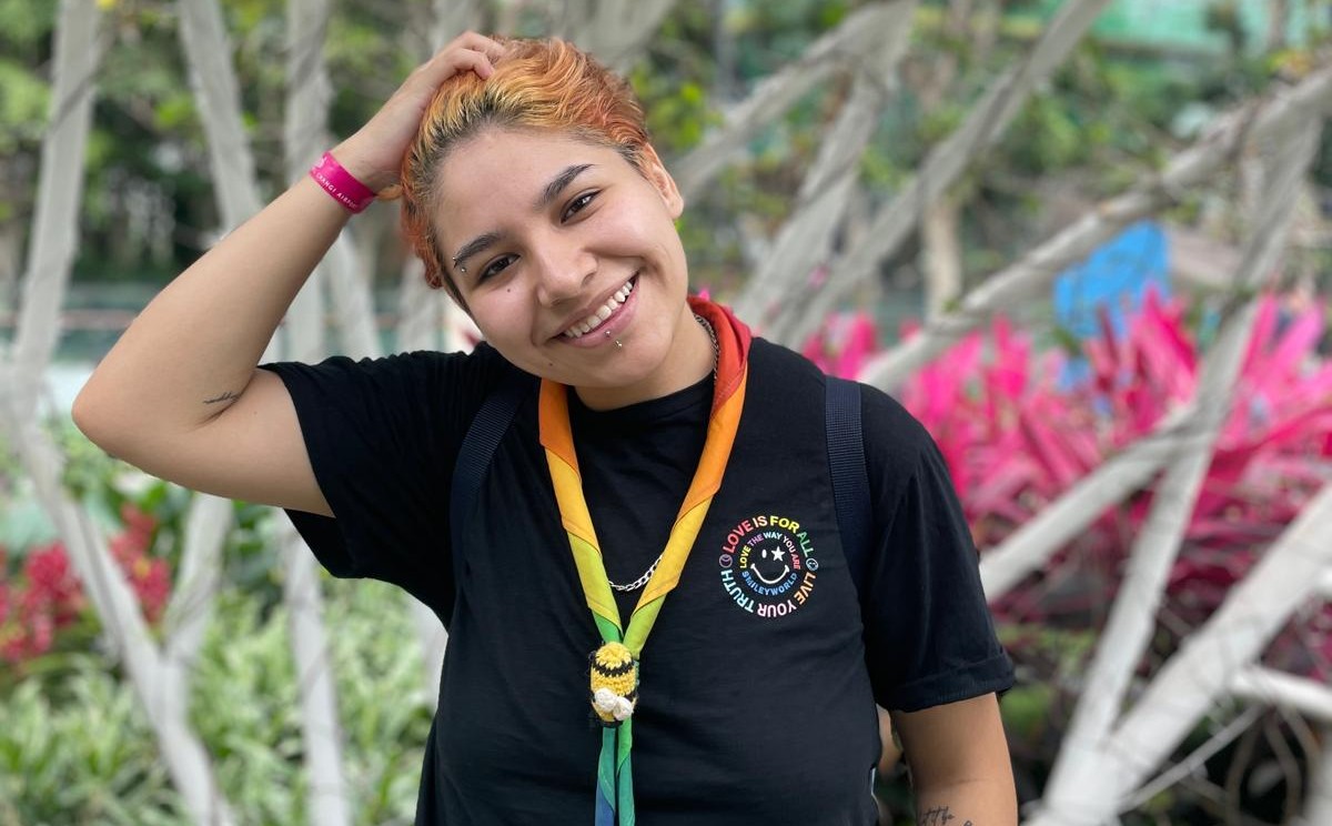Astro, a Scout volunteer wearing a rainbow necker, smiles at the camera while holding one hand in their hair.