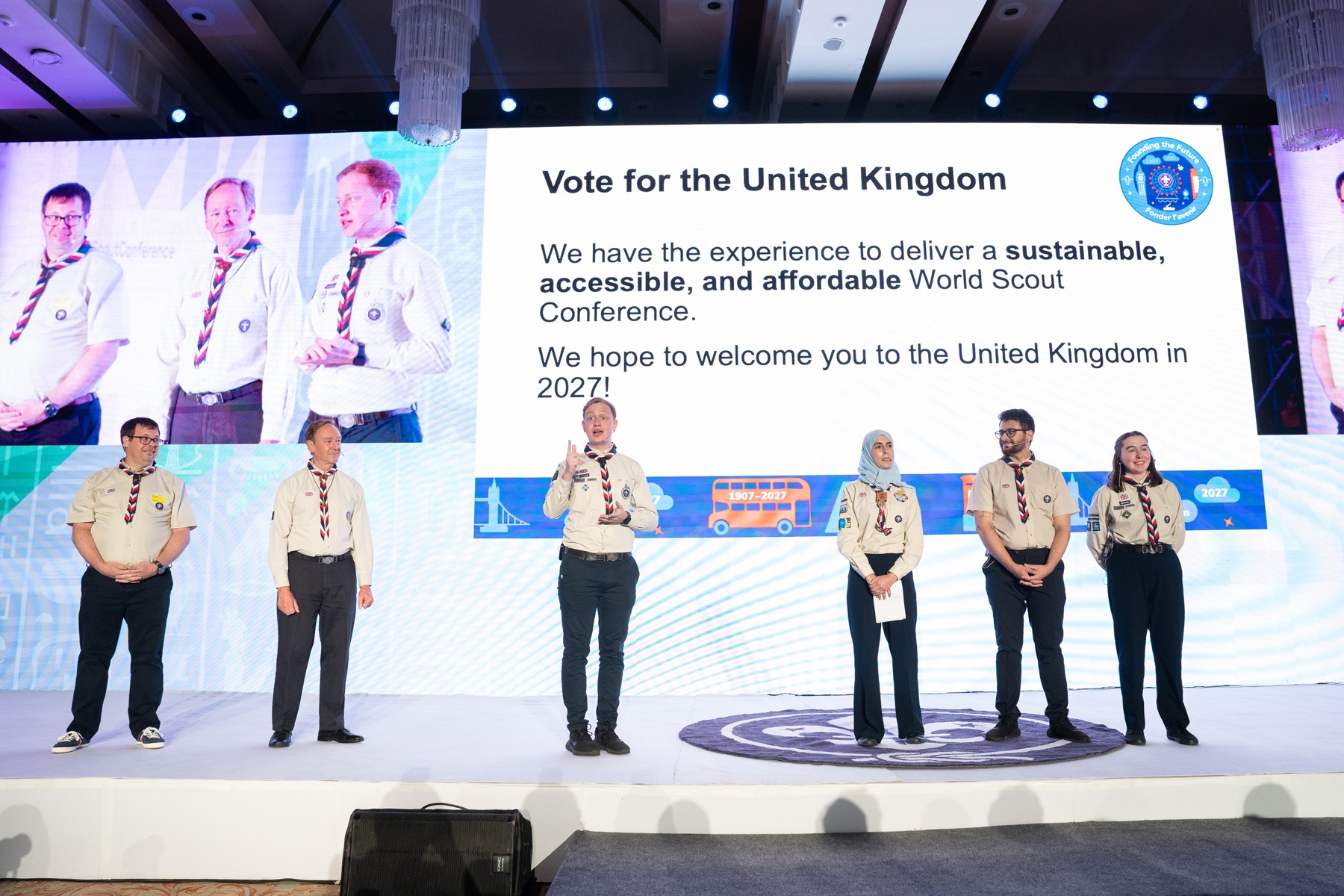 Five volunteers in UK Scout uniforms and neckers presenting their bid for hosting the World Scout Conference in 2027 on a stage.