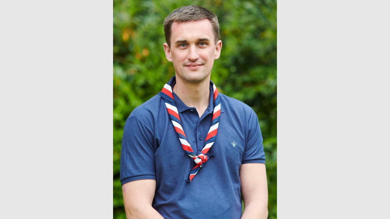 Callum is stood outside and in front of green leaves. He's looking at the camera with his arms by his sides, wearing a red, white and navy necker and a navy Scouts polo shirt.