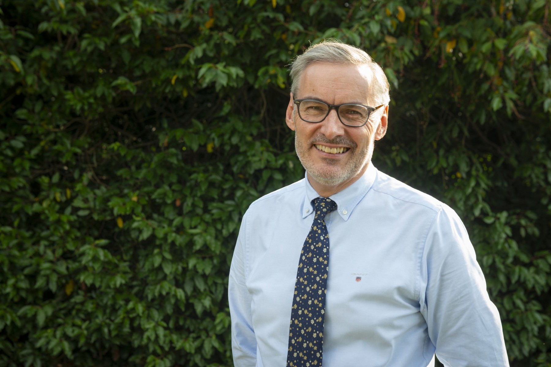 Aidan standing in front of a green bush, smiling and wearing glasses, a shirt and tie.