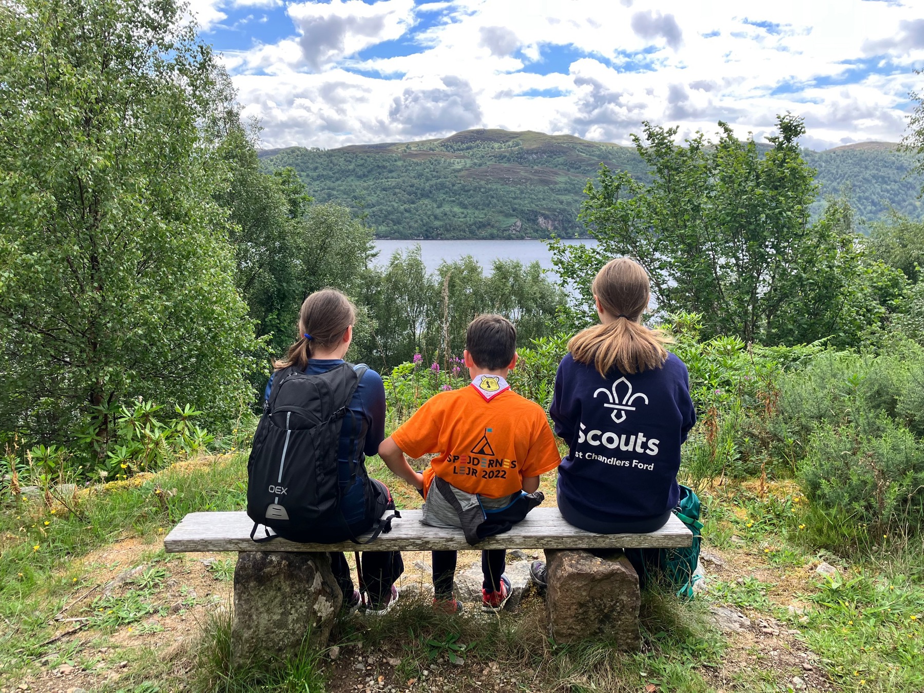 Hattie, Ollie and Bethan are sitting on a wooden bench surrounded by trees. We can see their backs as they look out at the view, with a lake in the distance.