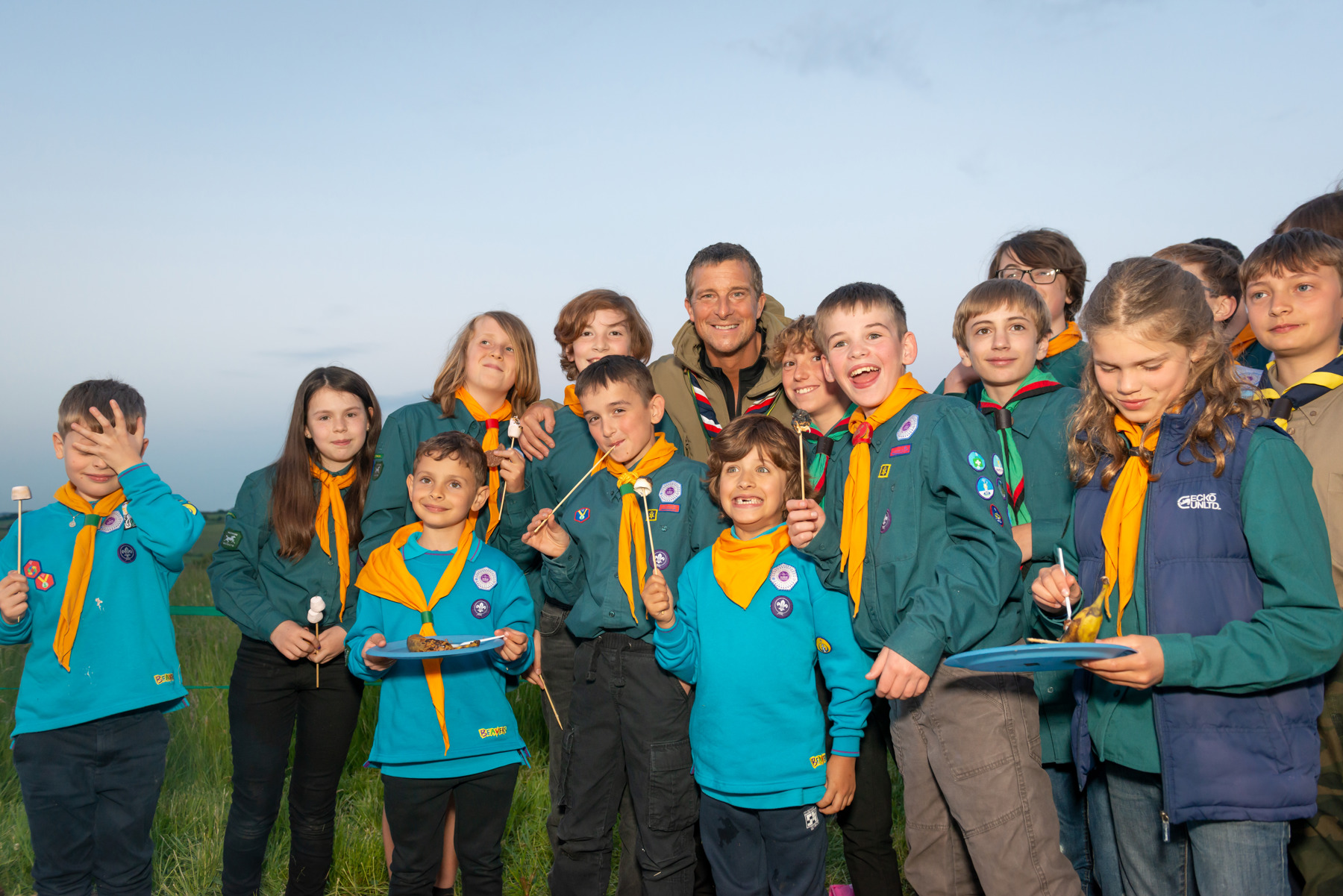 Chief Scout Bear Grylls is standing on grass with a group of Beavers and Cubs. All the Beavers and Cubs are in uniform with yellow neckers, and Bear Grylls is wearing a red navy and white necker. The sky behind them is a grey/blue.