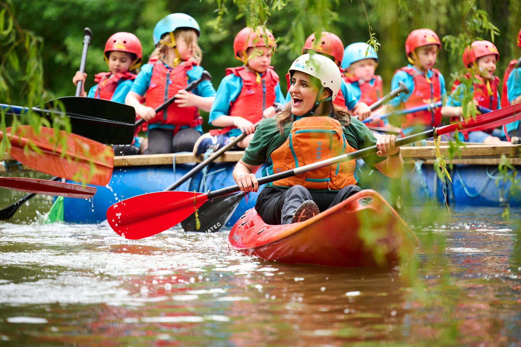 A smiling volunteer in a kayak paddles in front of rafts with young people wearing life jackets and helmets.