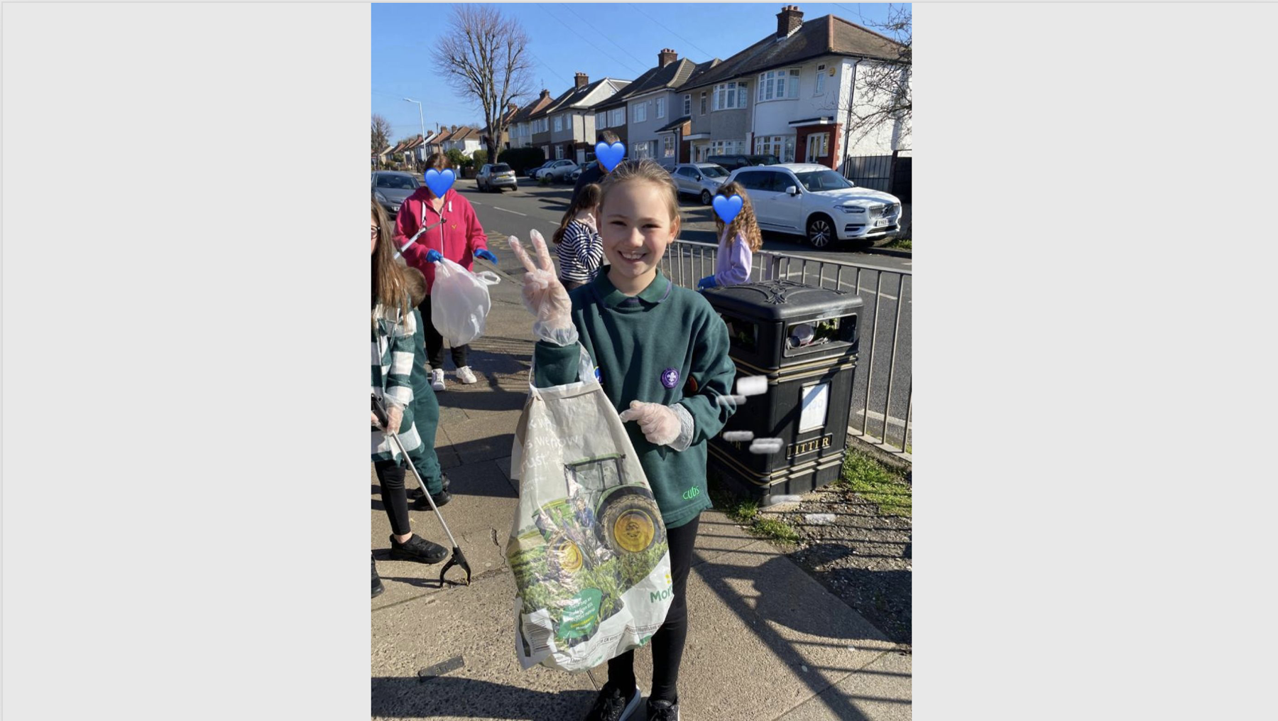 A Cub Scout is holding a carrier bag with gloves on. She's smiling at the camera in her Cubs uniform and doing a 'peace' sign with her right hand. She's standing on a pavement with a bin just behind her and there are other young people and adults in the background.
