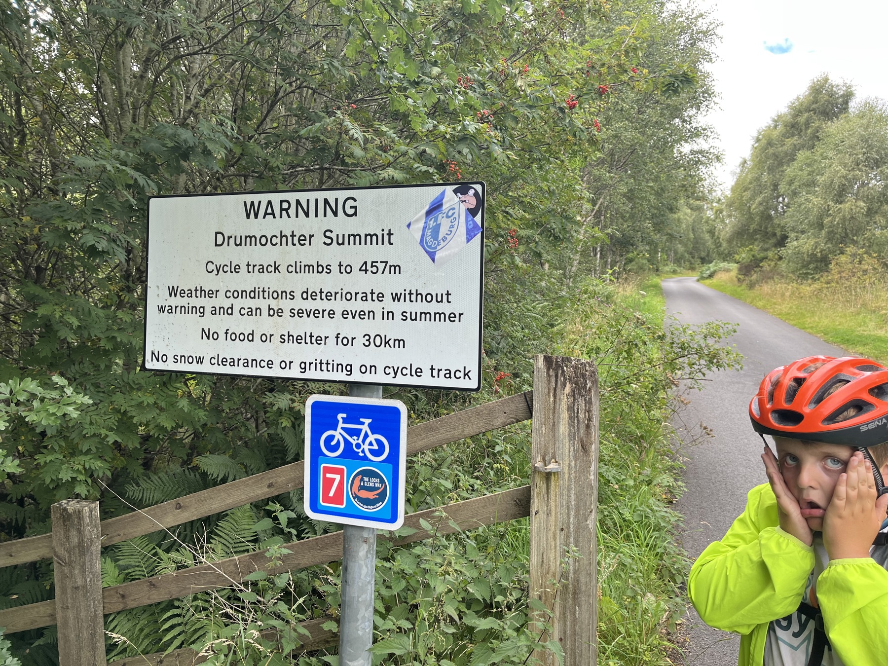 Harry with his hands to his face by a sign to Drumochter Summit that shows a cycle climb of 457 metres