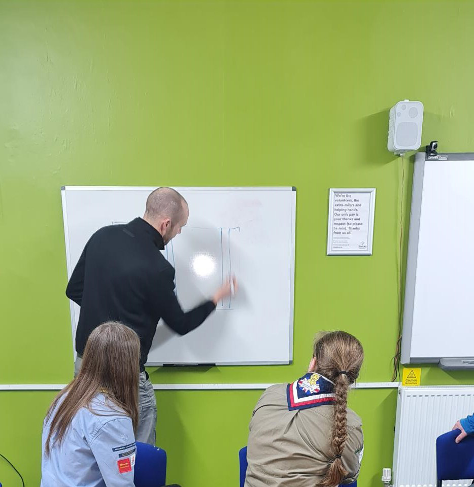 The image shows Merseyside-based artist, Paul Curtis, drawing on a whiteboard. The whiteboard is on a green wall, and there are a few Scouts facing him as he writes on the board. Together, they're coming up with ideas for the Wirral Scouts mural.