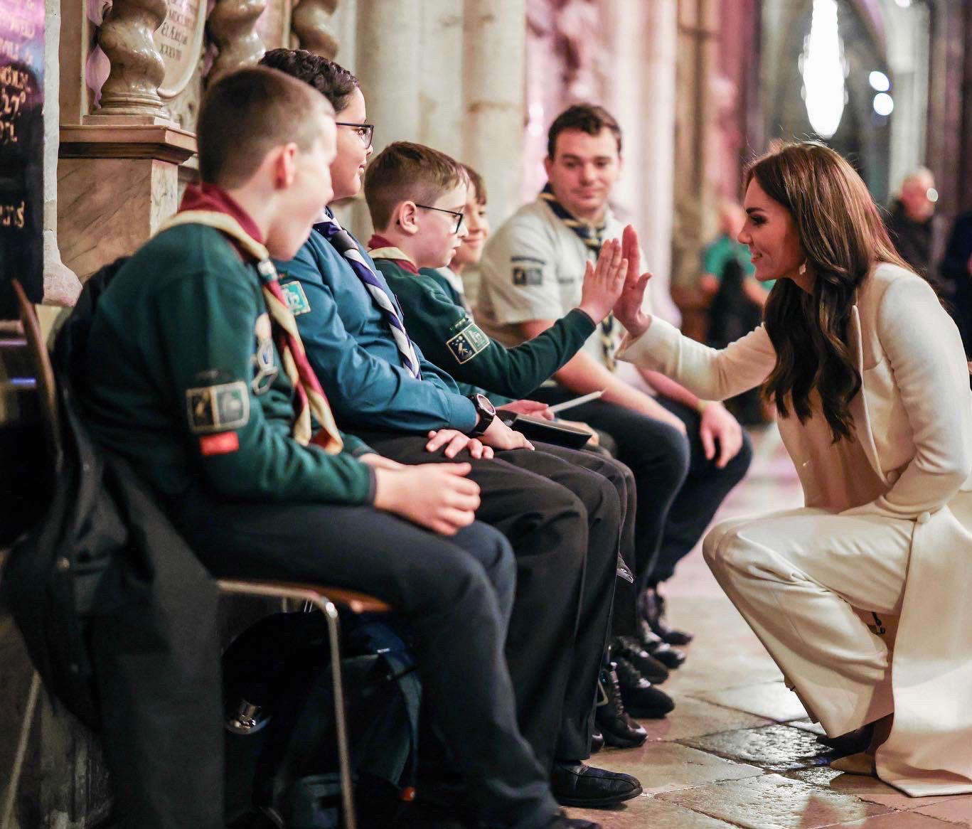 Three Cubs and a volunteer uniform and neckers are sat next to each other to the left of the image, with one Cub holding his hand out to the meet the hand of HRH Princess of Wales, who's crouching down and facing the Cubs. They're inside Westminster Abbey.