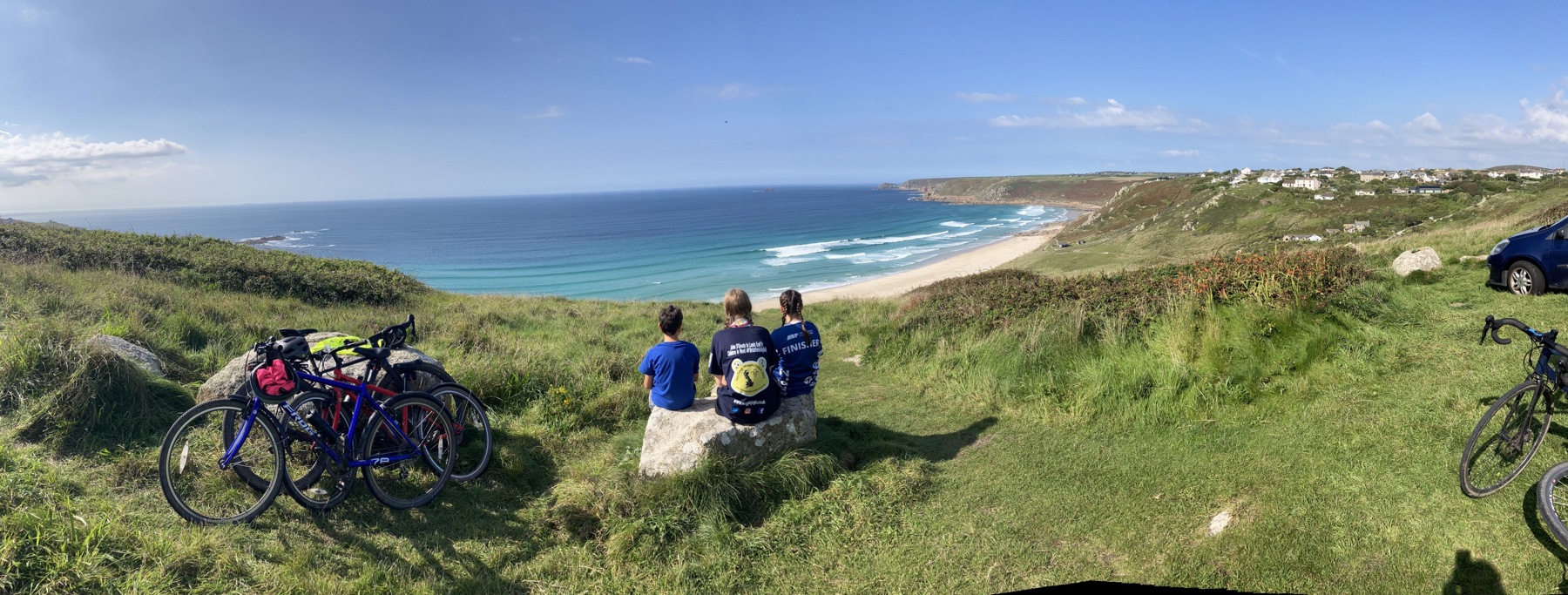 The image shows the backs of Ollie, Bethan and Hattie as they sit on a hill and look out towards the sea. Next to them are grassy hills and to their left are their bicycles and belongings. We can see the backs of their heads as they look at the view.