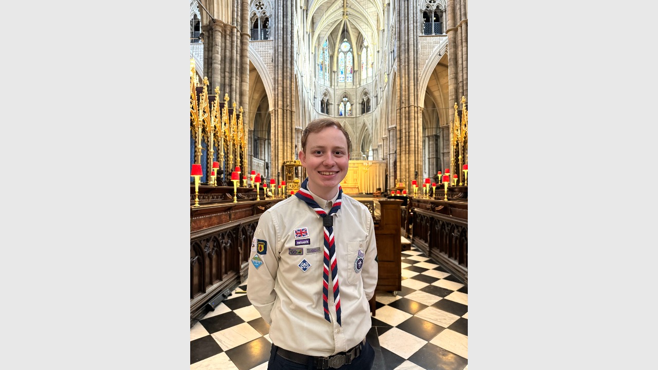The image shows UK Rep Pool member Luke wearing his Scout uniform and necker and smiling at the camera. He's standing inside a chapel with his arms behind his back