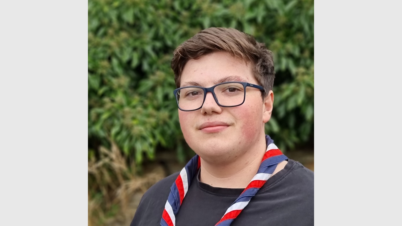 Alisdair is stood in front of a green hedge outside. He's wearing navy rectangular glasses, a red, white and navy necker and a navy top. He's smiling at the camera.