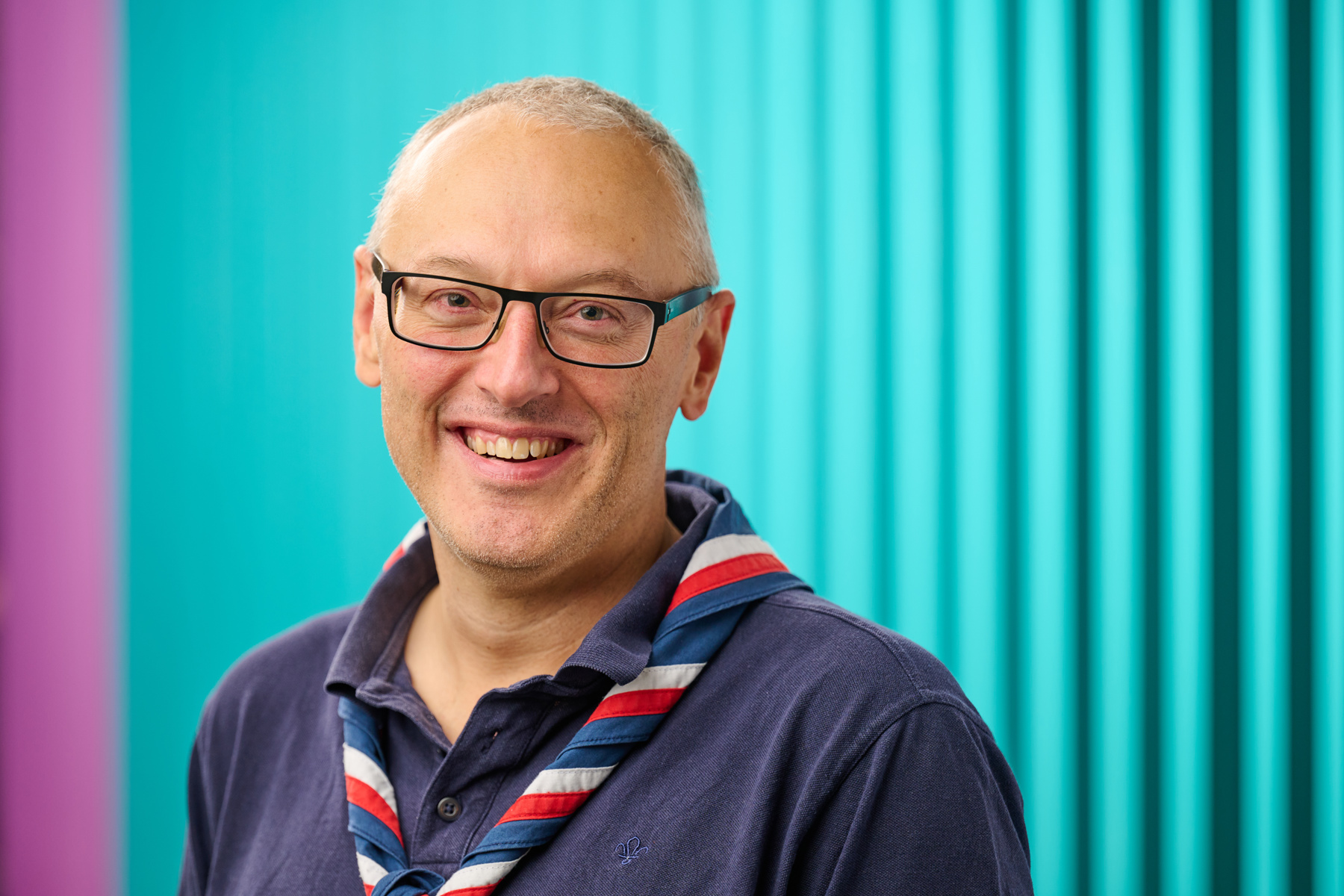 Nigel Ball smiling at the camera while wearing glasses, a navy Scouts polo shirt and stripy scarf