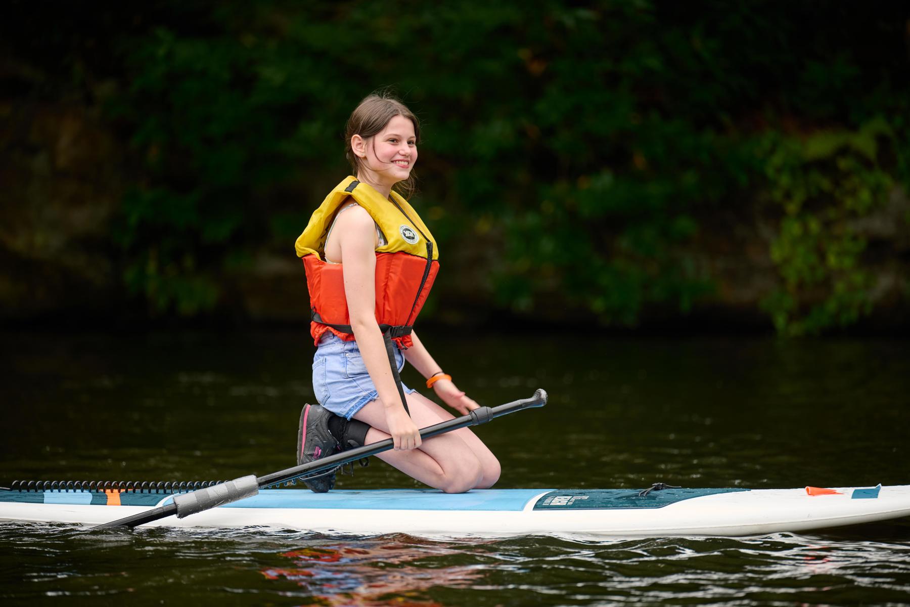 An Explorer knelt on a paddleboard on a lake. There's green trees and bushes in the background. She's wearing an orange and yellow life jacket, holding the paddle and looking to the right. She's smiling.