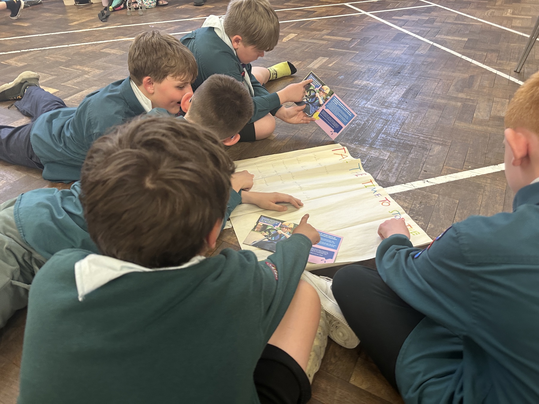 Five Cub Scouts are sat on the floor of a hall. They're holding copies of the Scout manifesto and have a piece of A3 paper in front of them as they do their Changemaker Challenge.