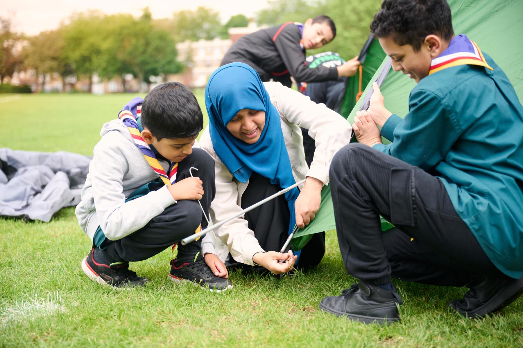 A volunteer wearing a blue hijab helps two Scouts pitch a tent outside.