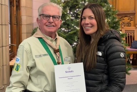 The image shows Kirsty Mack earning her 20 years of service award. She's stood with a black coat on, next to another volunteer who's on the left of the photo. They're both smiling at the camera as Kirsty holds her award certificate up.