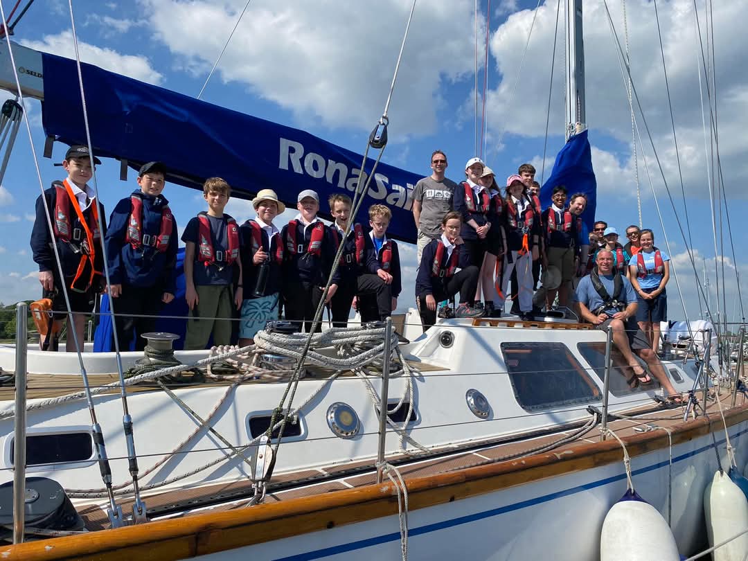 The image shows Scouts and volunteers stood on a boat. They're all wearing lifejackets and smiling at the camera, with the photo being taken off the boat. There is a banner saying 'Rona sail' behind them.