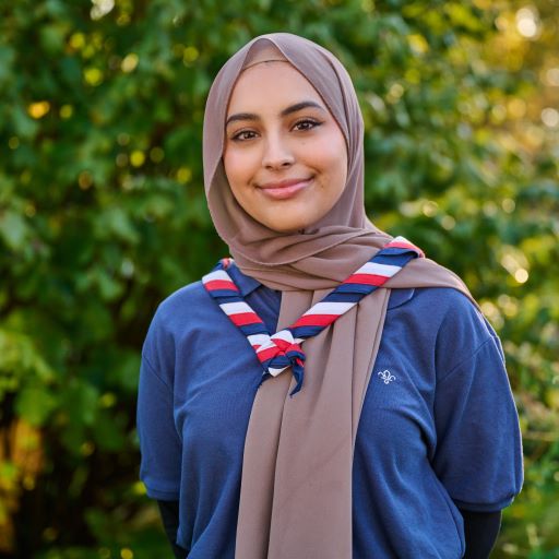 Ayesha is stood in front of trees. She is smiling and is wearing her hijab and Scouts necker. She looks relaxed and is wearing a blue Scouts top, with her hands behind her back.
