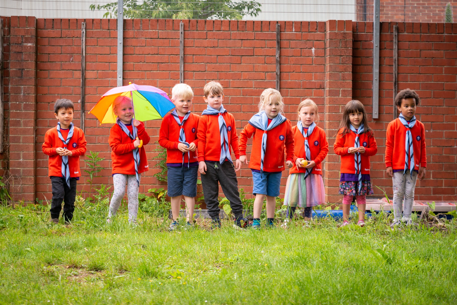 The image shows a group of Squirrels in uniform in front of a brick wall and standing on grass. They're all stood in a line with one Squirrel to the left holding a multicoloured umbrella.