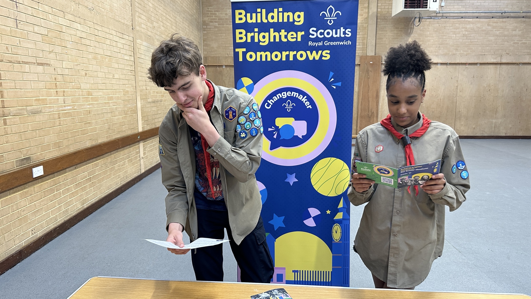 Two Scouts are stood in a hall in front of a table. They're both in uniform and reading the Changemaker Challenge and Scout manifesto. They're stood in front of a standing banner which says 'Building brighter tomorrows' to promote the Changemaker Challenge and badge. 