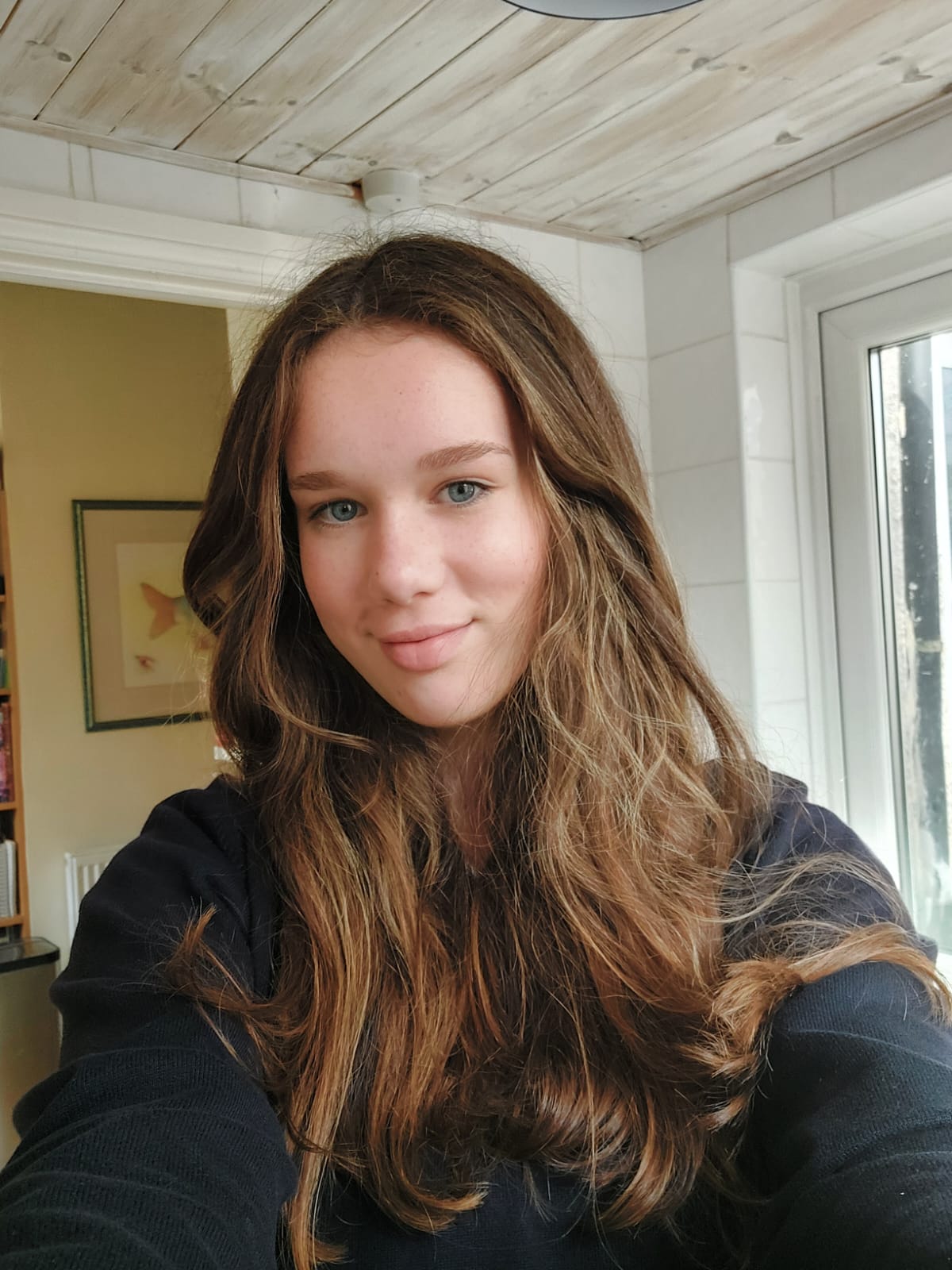 This image shows Hannah, a teenage girl with long brown hair who's smiling at the camera. She's taking the selfie in a room with tiled walls. She wears a long sleeved black jumper.