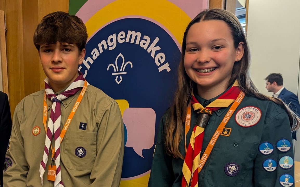 Two young people smile at the camera while standing in front of a graphic of the changemaker badge.