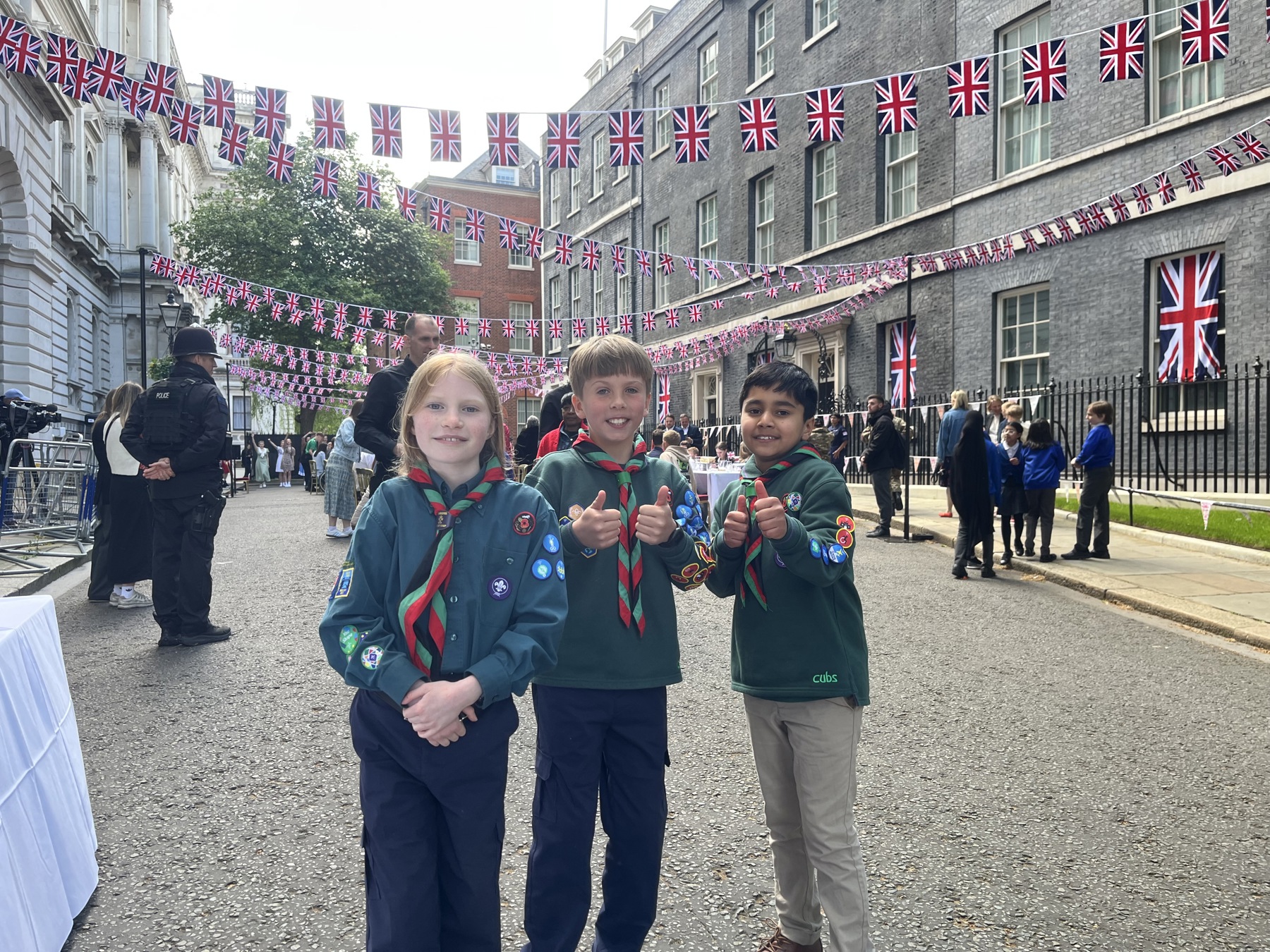 The image shows three Cubs on Downing Street celebrating VE Day 80. The three Cubs are all wearing their uniform and neckers. There are Union Jack flags on bunting going down the street with crowds of people behind them and tables laid out for a street party.